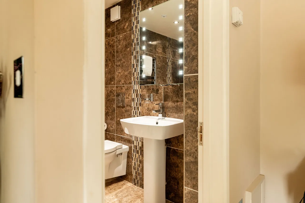 Modern bathroom with brown marble tiles, a pedestal sink, wall-mounted toilet, and an illuminated mirror above the sink.