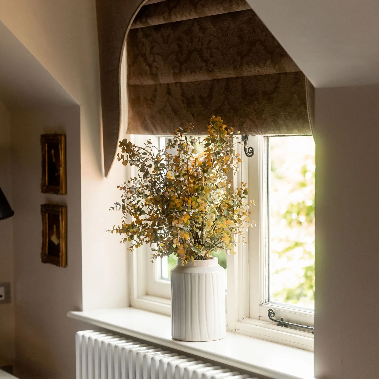 A vase with yellow flowers placed on a windowsill in front of a window with brown roman shades, above a white radiator.