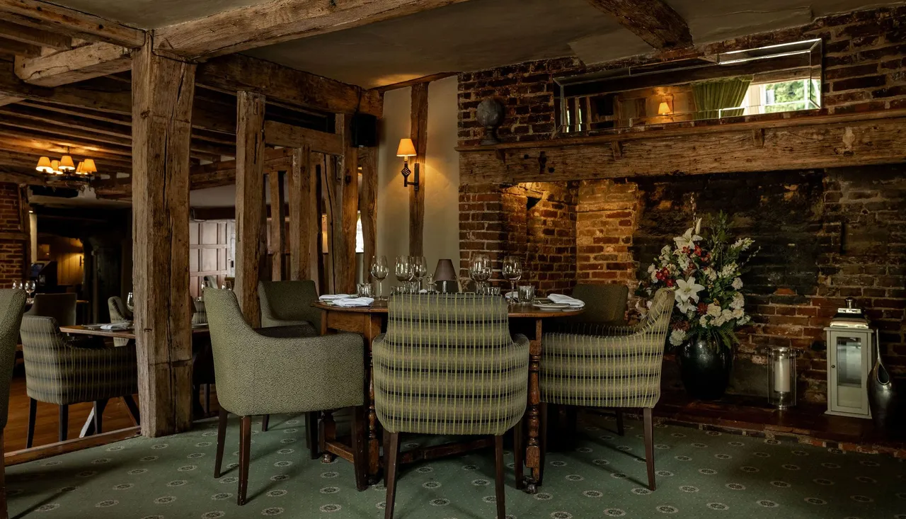 Cozy rustic restaurant interior with wooden beams, brick walls, green upholstered chairs, set dining tables, floral arrangement, and soft lighting.