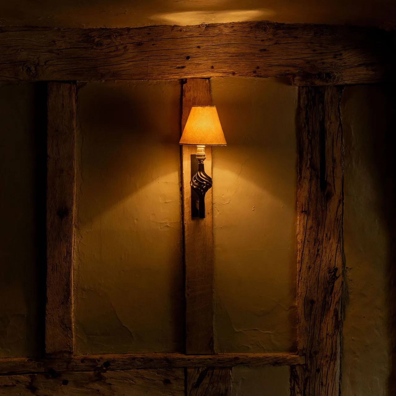 A warmly lit wall sconce with a yellow lampshade mounted on a rustic, wooden beam wall in a dimly lit room.