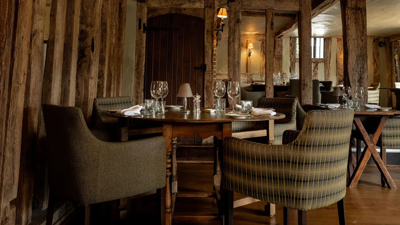 Rustic restaurant interior with wooden beams, cozy upholstered chairs, and neatly set tables with glassware and napkins.