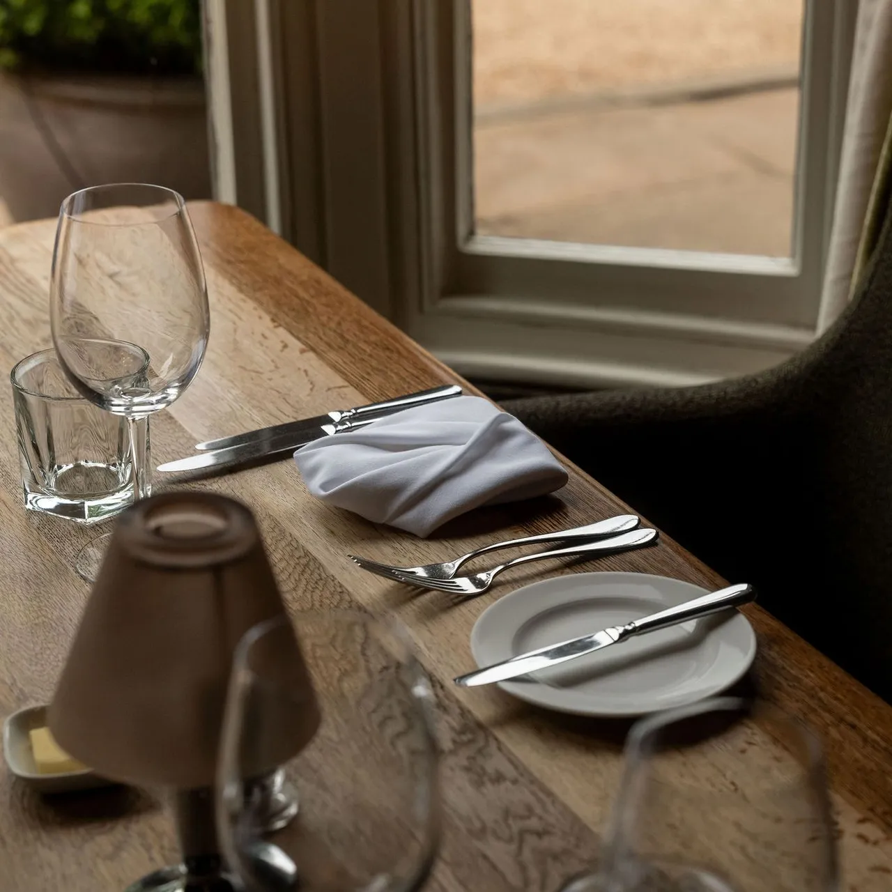 A close-up of a neatly set dining table by a window, featuring a wine glass, water glass, silverware, a plate with a butter knife, a folded white napkin, and a small lamp.