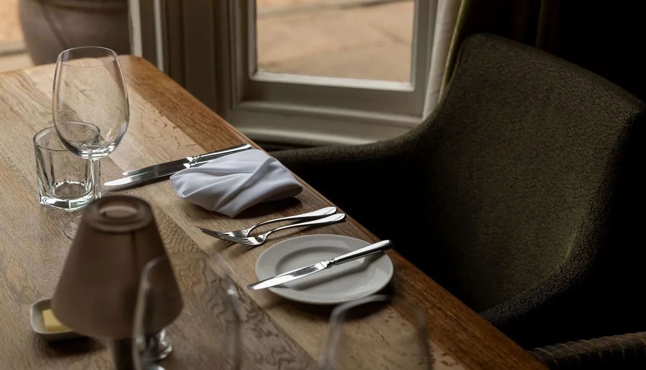 A close-up of a neatly set dining table by a window, featuring a wine glass, water glass, silverware, a plate with a butter knife, a folded white napkin, and a small lamp.