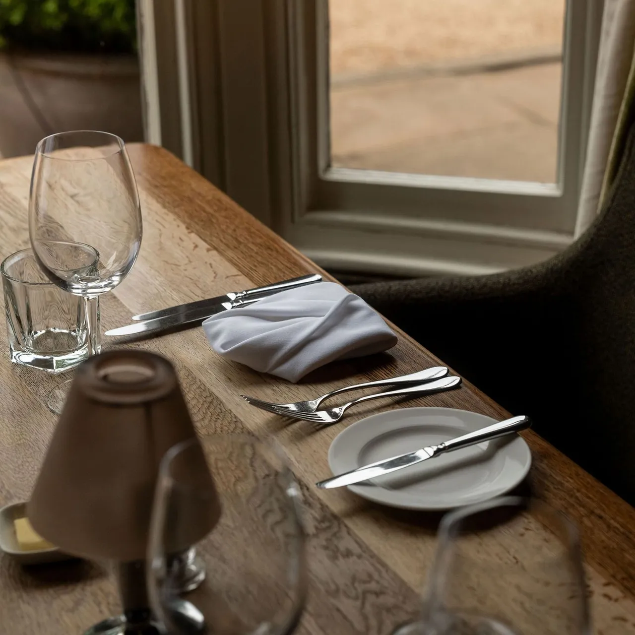 A close-up of a neatly set dining table by a window, featuring a wine glass, water glass, silverware, a plate with a butter knife, a folded white napkin, and a small lamp.