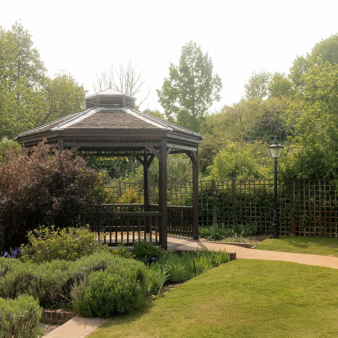 Wooden gazebo in a landscaped garden surrounded by green bushes, flowers, and trees with a pathway and a lamppost nearby.