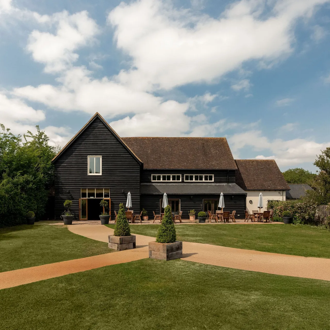 A large black wooden barn with a sloped roof, outdoor seating with tables and umbrellas, surrounded by well-kept grass, shrubbery, and a clear sky with scattered clouds.