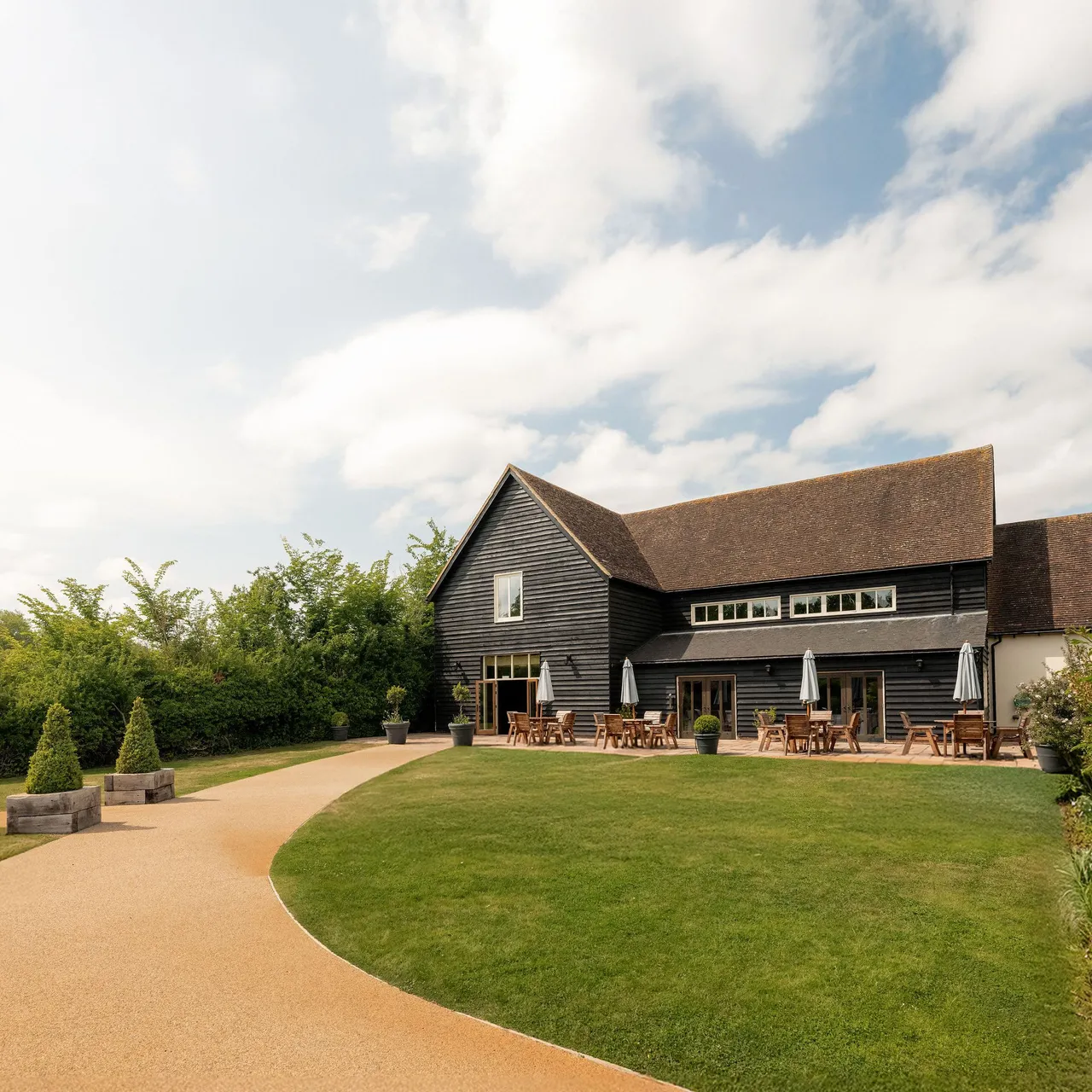 A modern barn-style building with outdoor seating, surrounded by green lawn and a curved pathway under a partly cloudy sky.