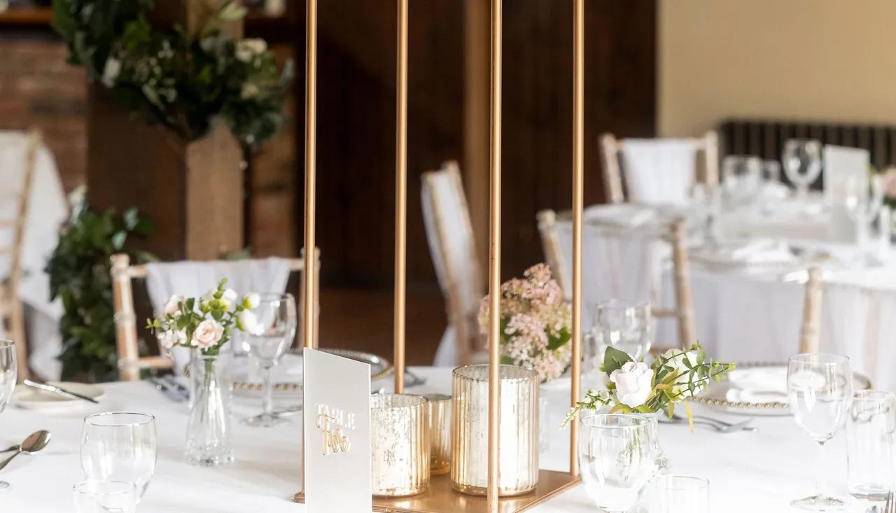 Elegant wedding table centerpiece featuring a tall arrangement of white and blush flowers with greenery on a gold stand, surrounded by glassware and candles in a decorated reception venue.