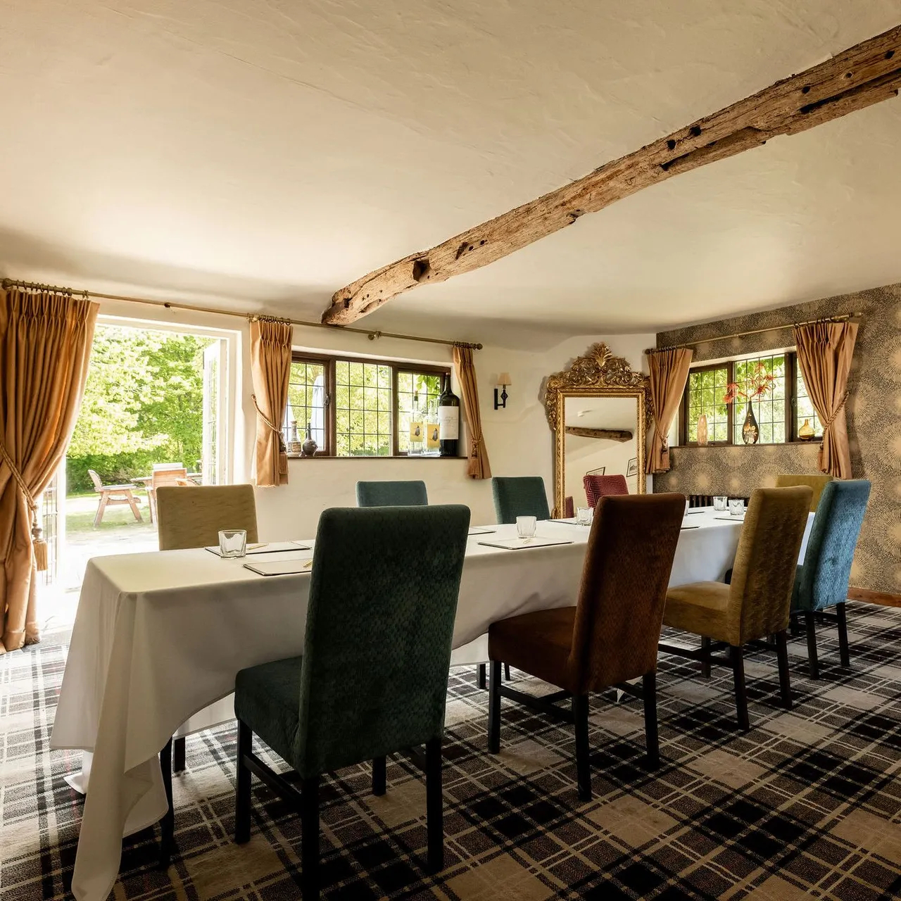 Elegant dining or meeting room with a set table and colorful chairs, featuring rustic wooden beams, patterned wallpaper, and large windows with gold curtains overlooking a garden.