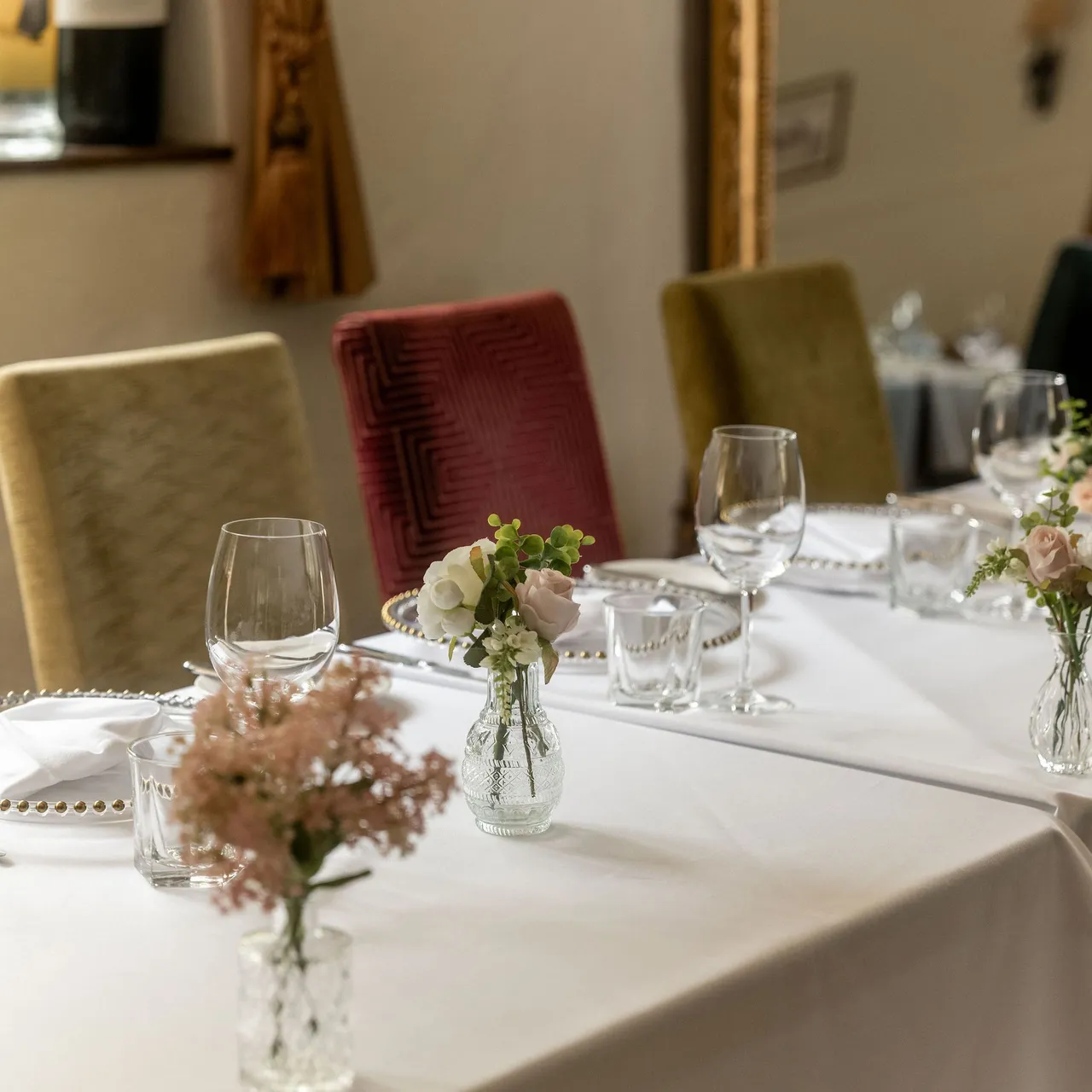 Elegant dining room setup with a white tablecloth, glassware, small floral vases, and upholstered chairs beside a large ornate mirror.