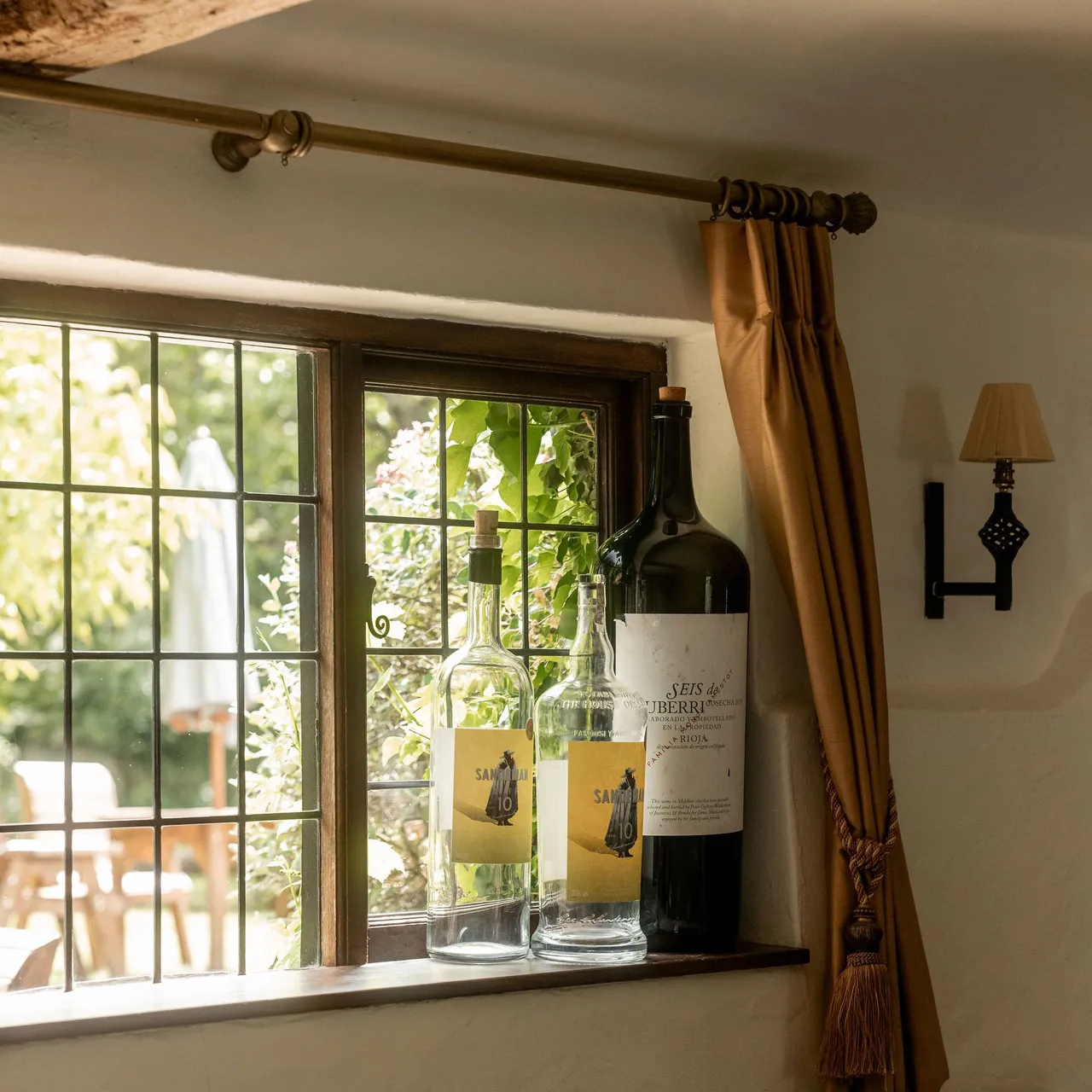 Three large wine bottles on a windowsill with a garden view through a leaded glass window in a rustic room.