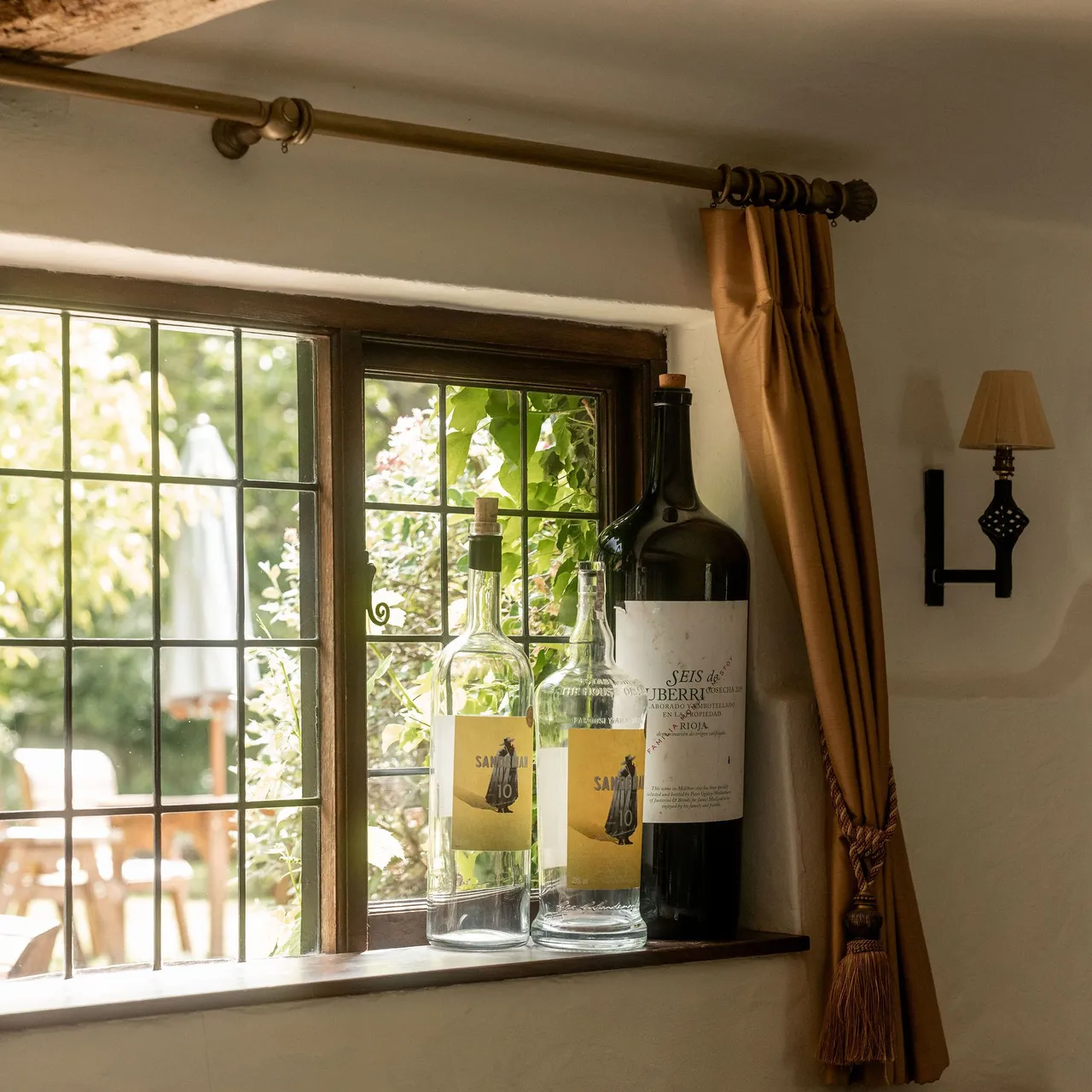 Three large wine bottles on a windowsill with a garden view through a leaded glass window in a rustic room.