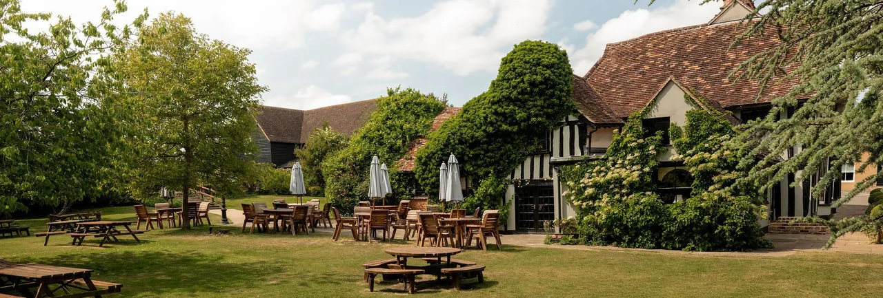 Outdoor seating area with wooden tables and chairs on a grassy lawn in front of a rustic building covered in green ivy.