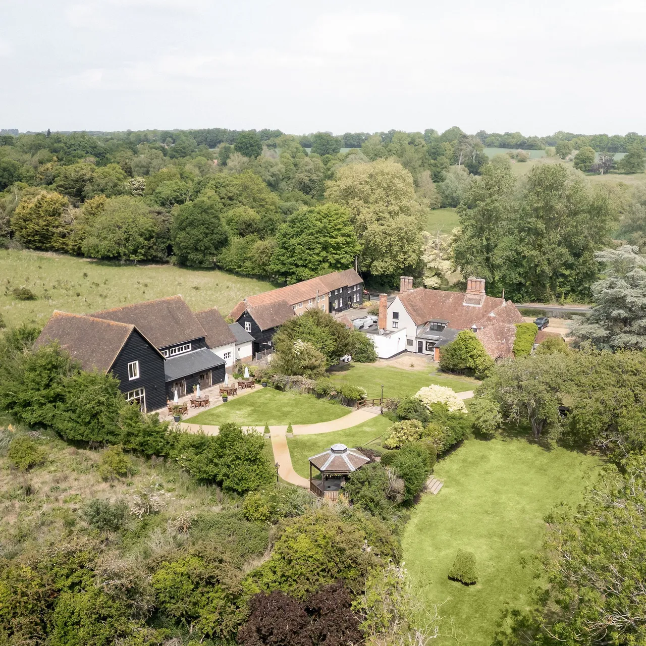 Aerial view of a countryside estate with several buildings, a gazebo, gardens, and large grassy areas surrounded by trees.