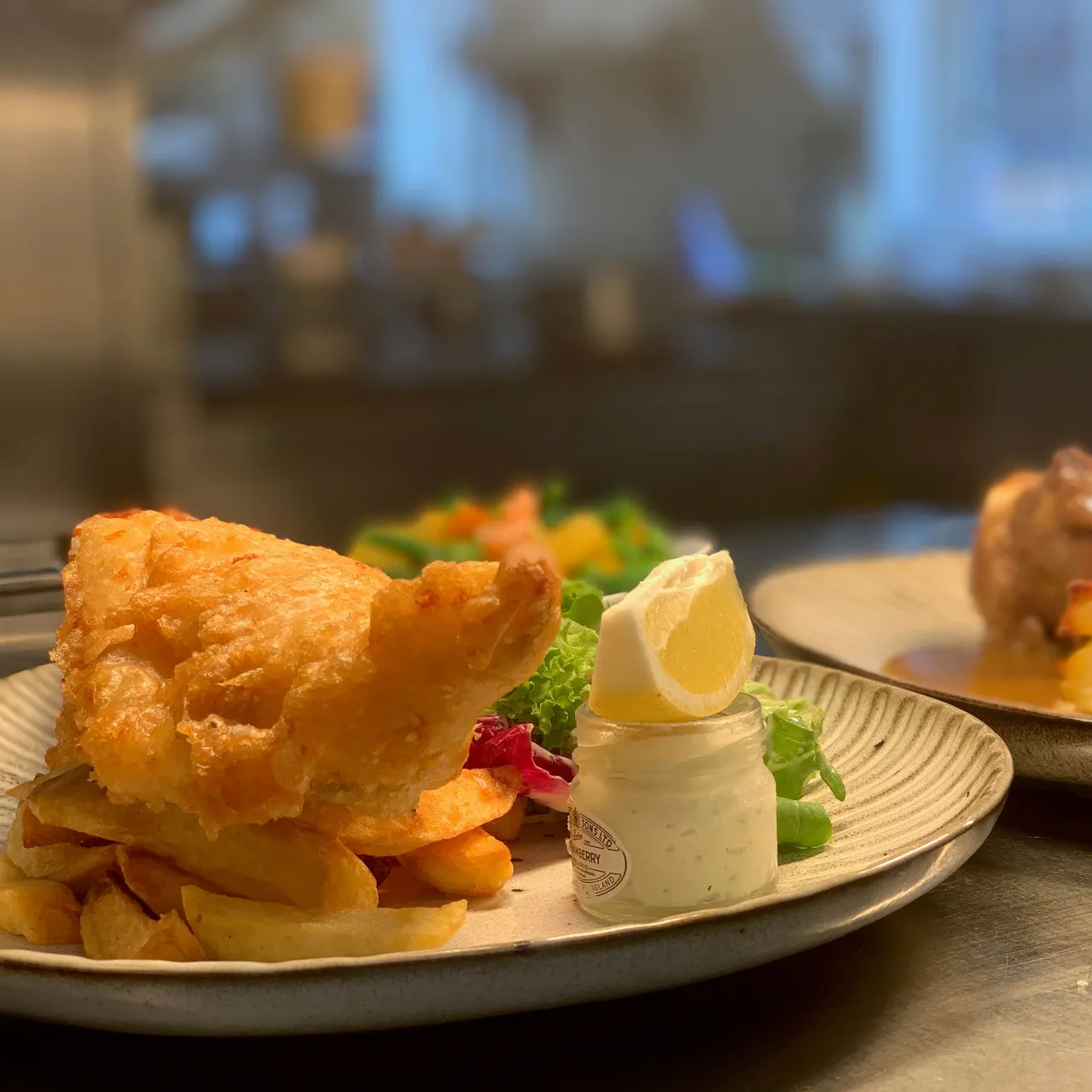 A plate of fish and chips with a jar of tartar sauce and lemon wedge, alongside another plate with layered potatoes and meat in the background.