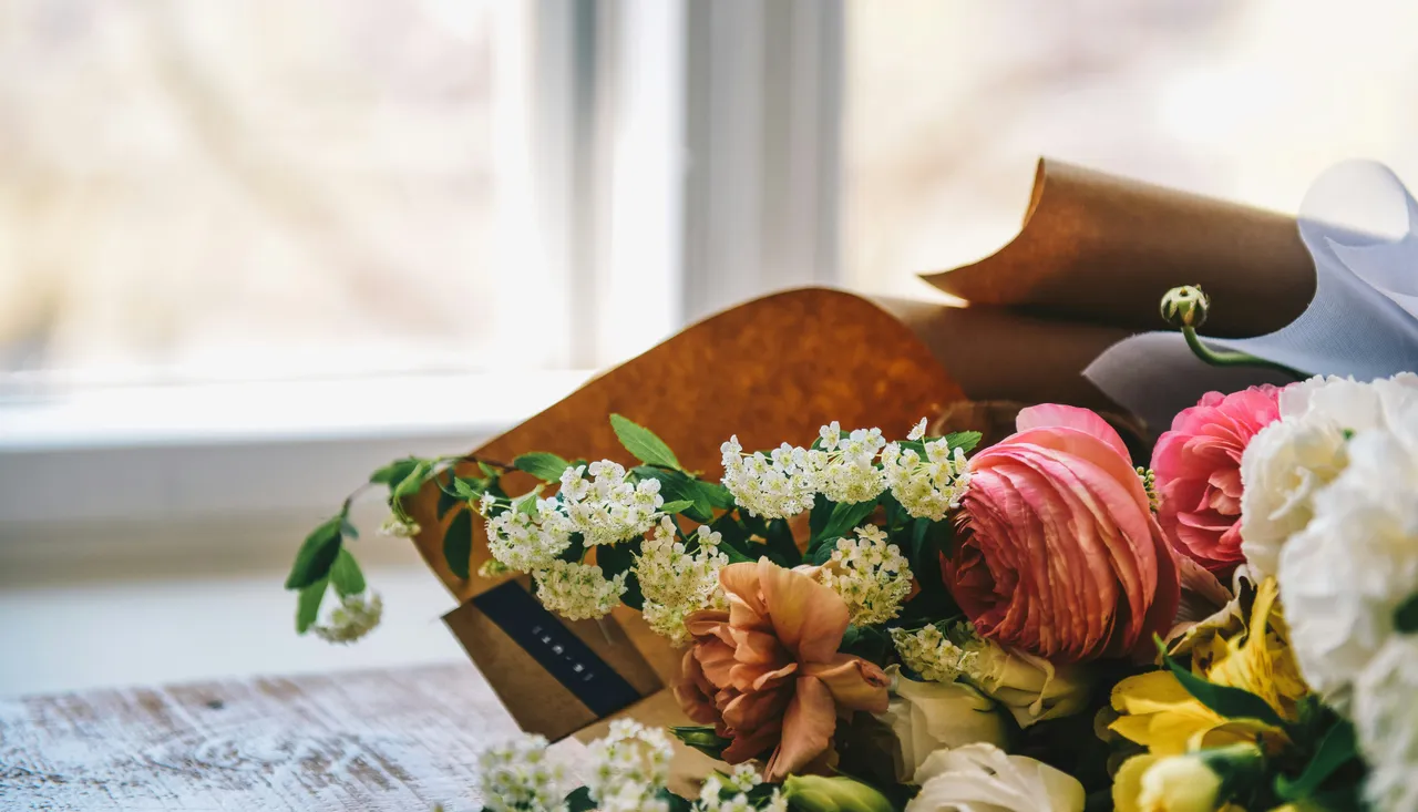 A close-up of a colorful bouquet of flowers, including pink ranunculus and white blossoms, placed on a wooden table near a window.