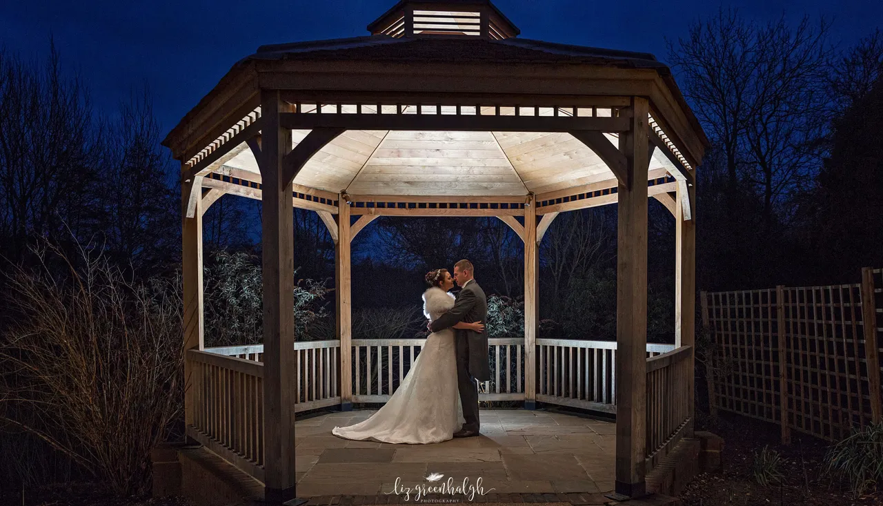 Bride and groom embrace in a beautifully lit wooden gazebo at night.
