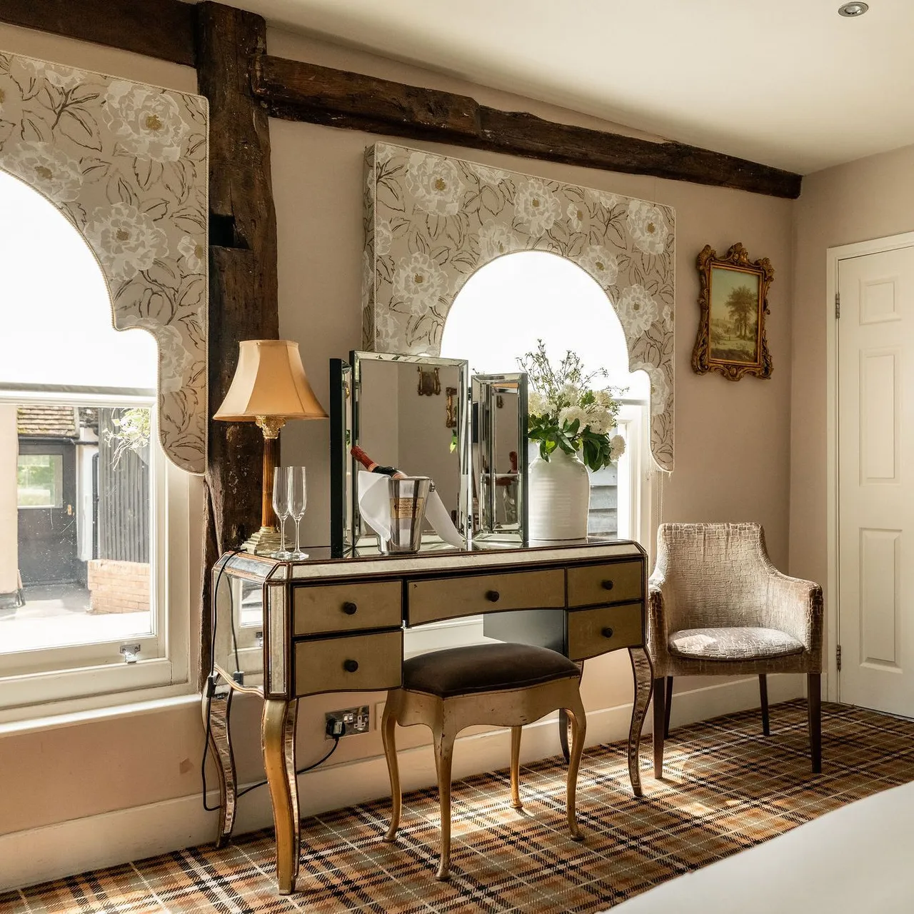 Elegant hotel room with vintage decor, including a mirrored vanity table, chair, lamp, framed art, flowers in a vase, wall-mounted TV, and patterned carpet.