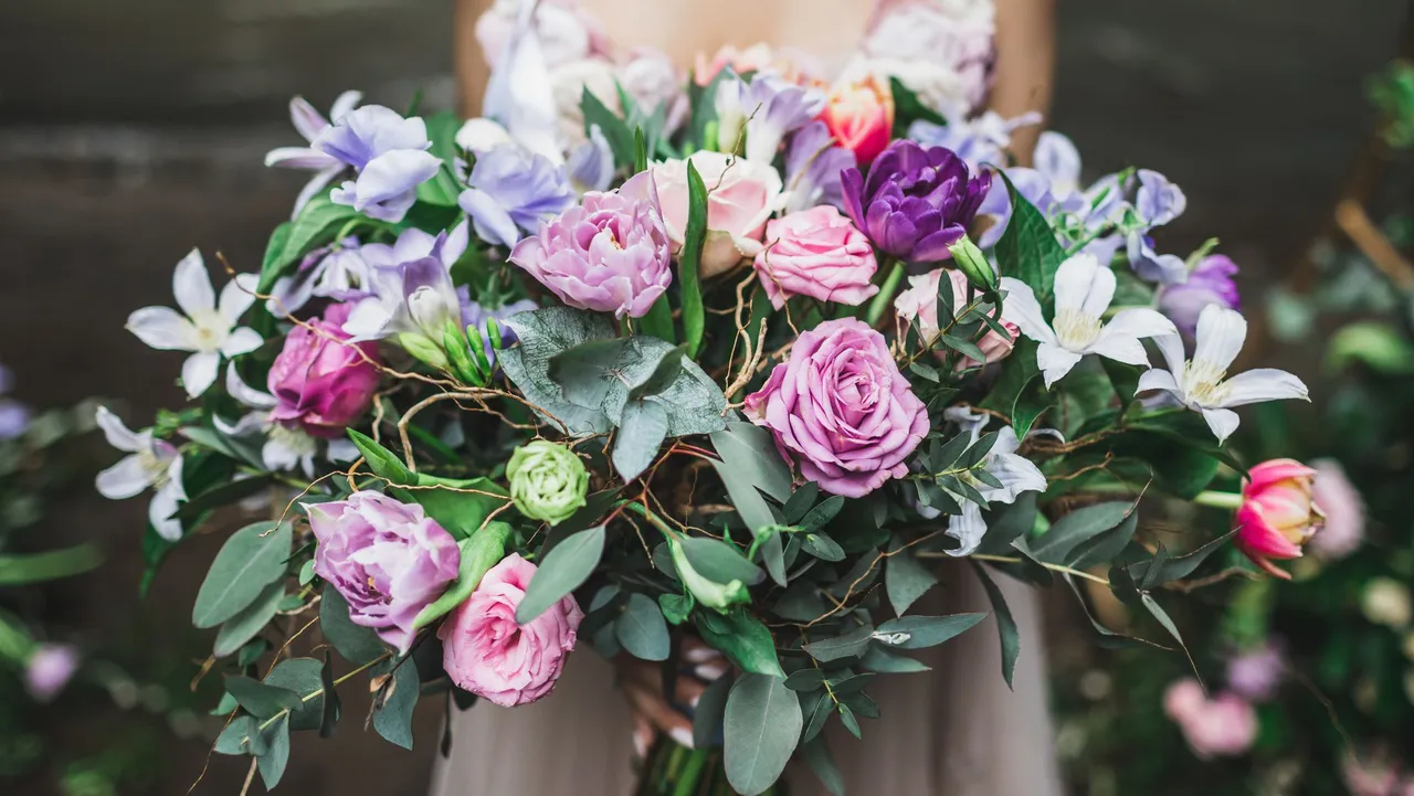 A bride holds a lush bouquet of roses, tulips, and wildflowers in shades of pink, purple, and white, accented with greenery and delicate vines.