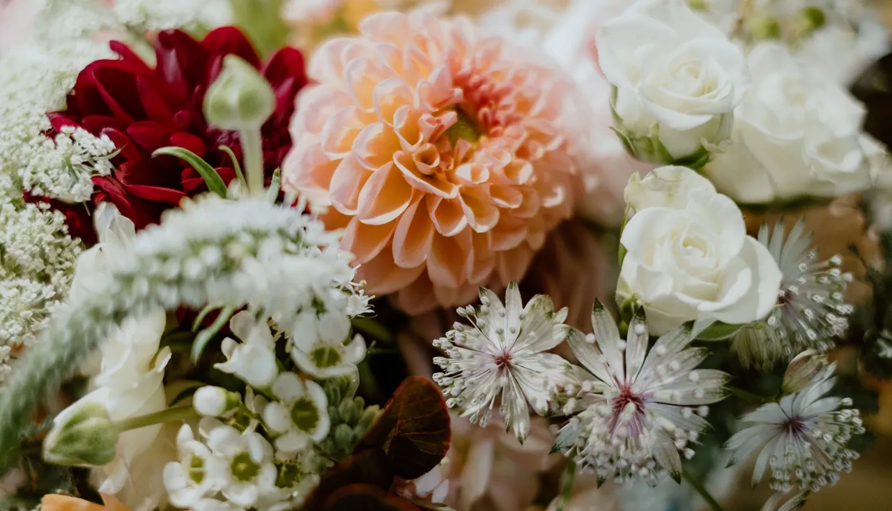 A close-up of a floral bouquet featuring a peach dahlia, white roses, deep red blooms, and delicate wildflowers, with lush greenery adding texture.