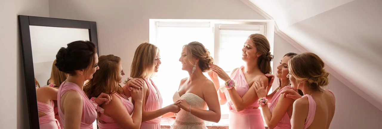 Bride in a white gown smiles surrounded by six bridesmaids in matching pink dresses, helping her get ready in a softly lit room with a sloped ceiling.