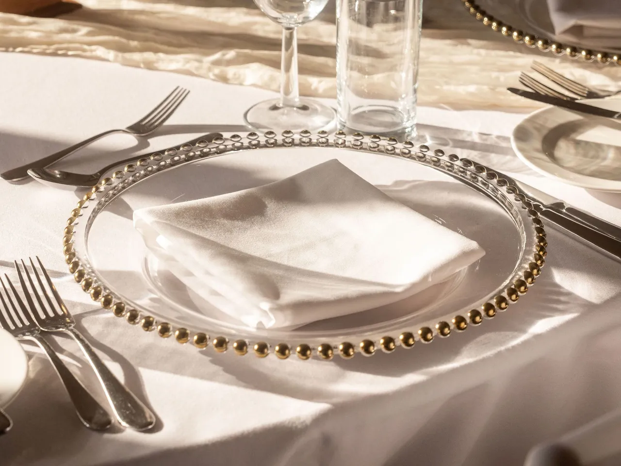Elegant table setting with glassware, silverware, and gold-beaded charger plates on a white tablecloth.