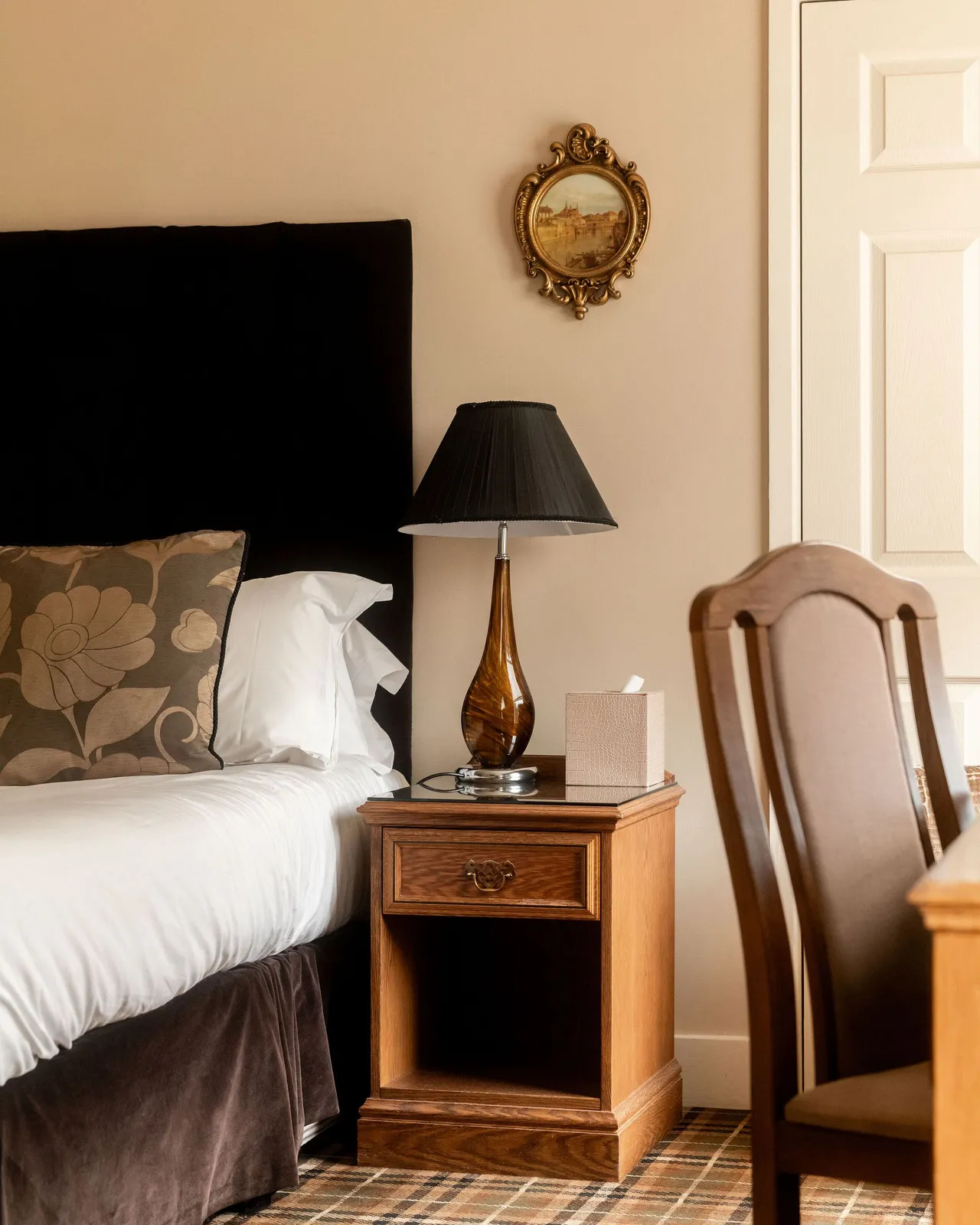 Cozy bedroom corner with a bed, floral pillow, wooden nightstand, brown table lamp, tissue box, and ornate framed painting on the wall.