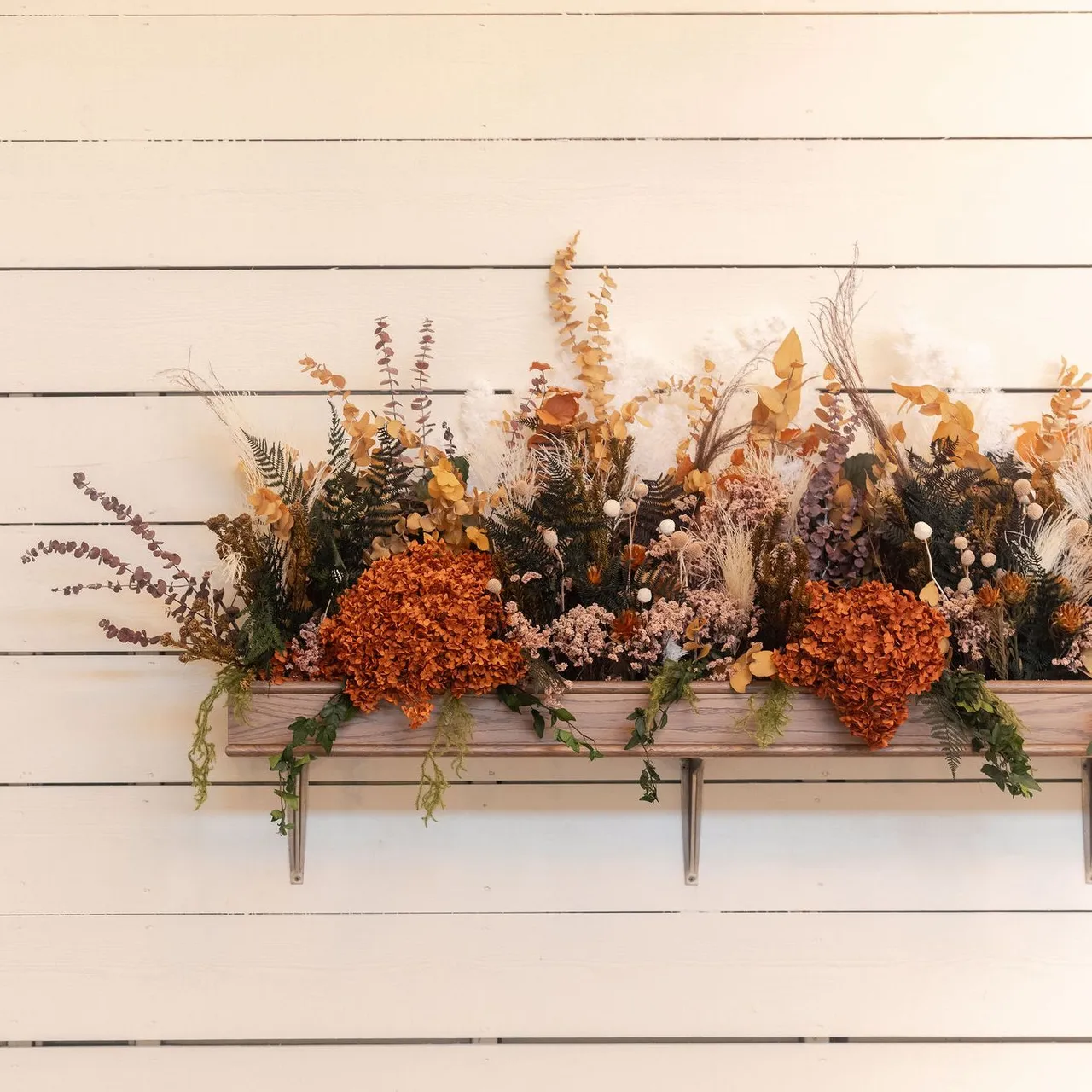 Wall-mounted wooden planter box filled with dried flowers and autumnal foliage against a white shiplap wall.
