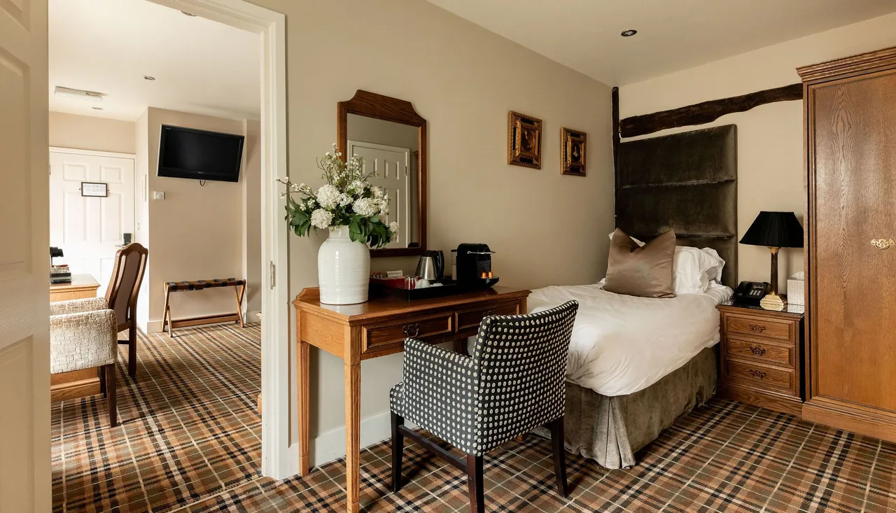 Elegant hotel room with plaid carpet, single bed, wooden furniture, desk with a vase of flowers, wall art, and a flat-screen TV in an adjacent sitting area.