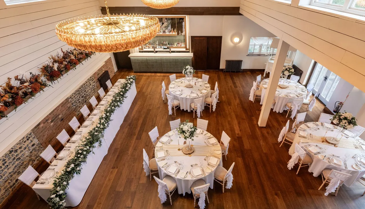 Elegant dining hall set up for an event with round tables, a long rectangular table, white chairs, and floral centerpieces.