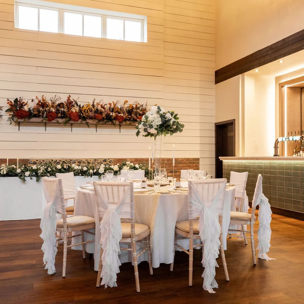 Elegant dining room with round table set for an event, featuring white chair covers and floral centerpiece, with a bar in the background.