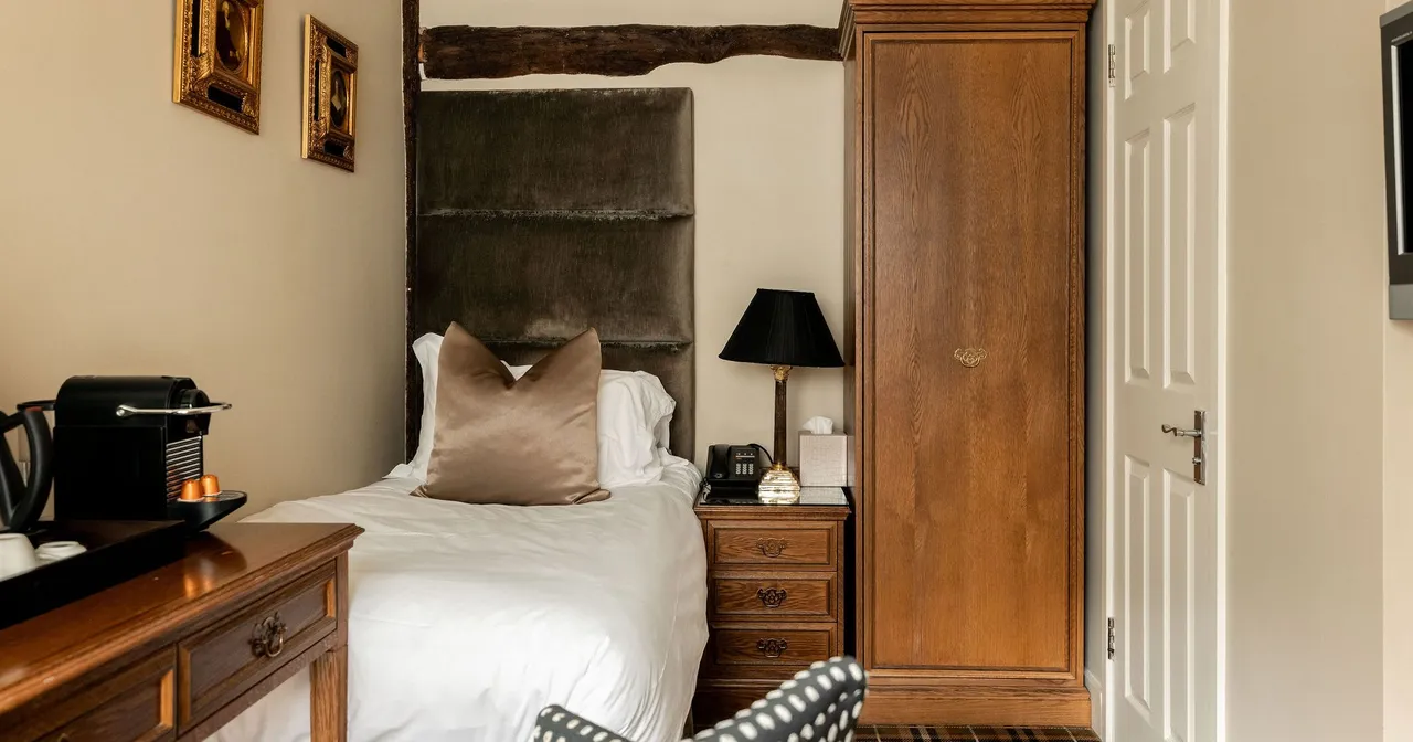 Cozy, traditionally furnished single bedroom with a neatly made bed, wooden furniture, a desk with a coffee machine, decorative pillows, wall art, and a checked carpet.