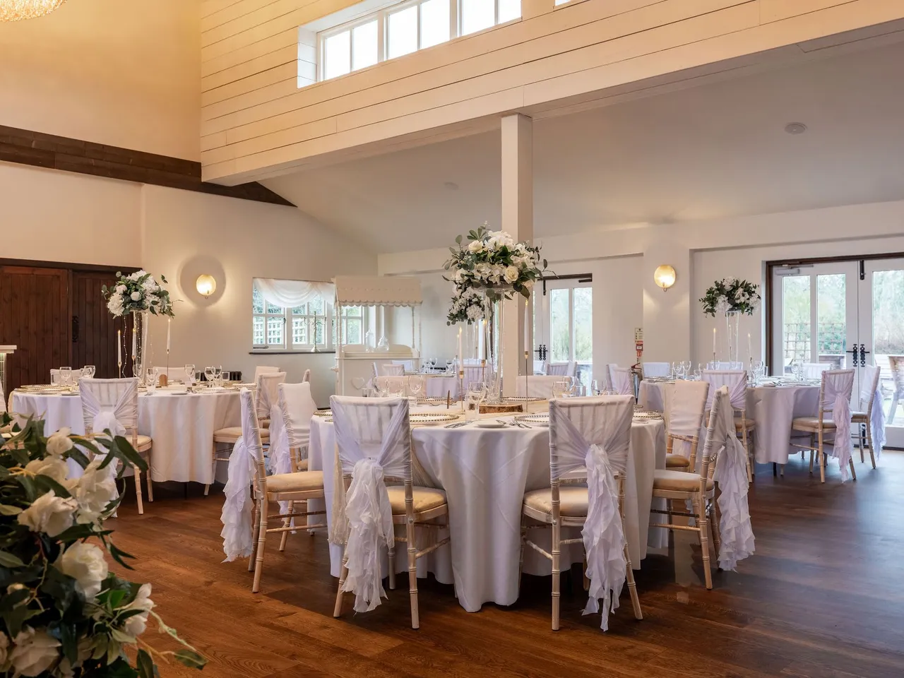 Elegant wedding reception dining room with round tables draped in white linens, decorated with tall floral centerpieces and chairs tied with white sashes.