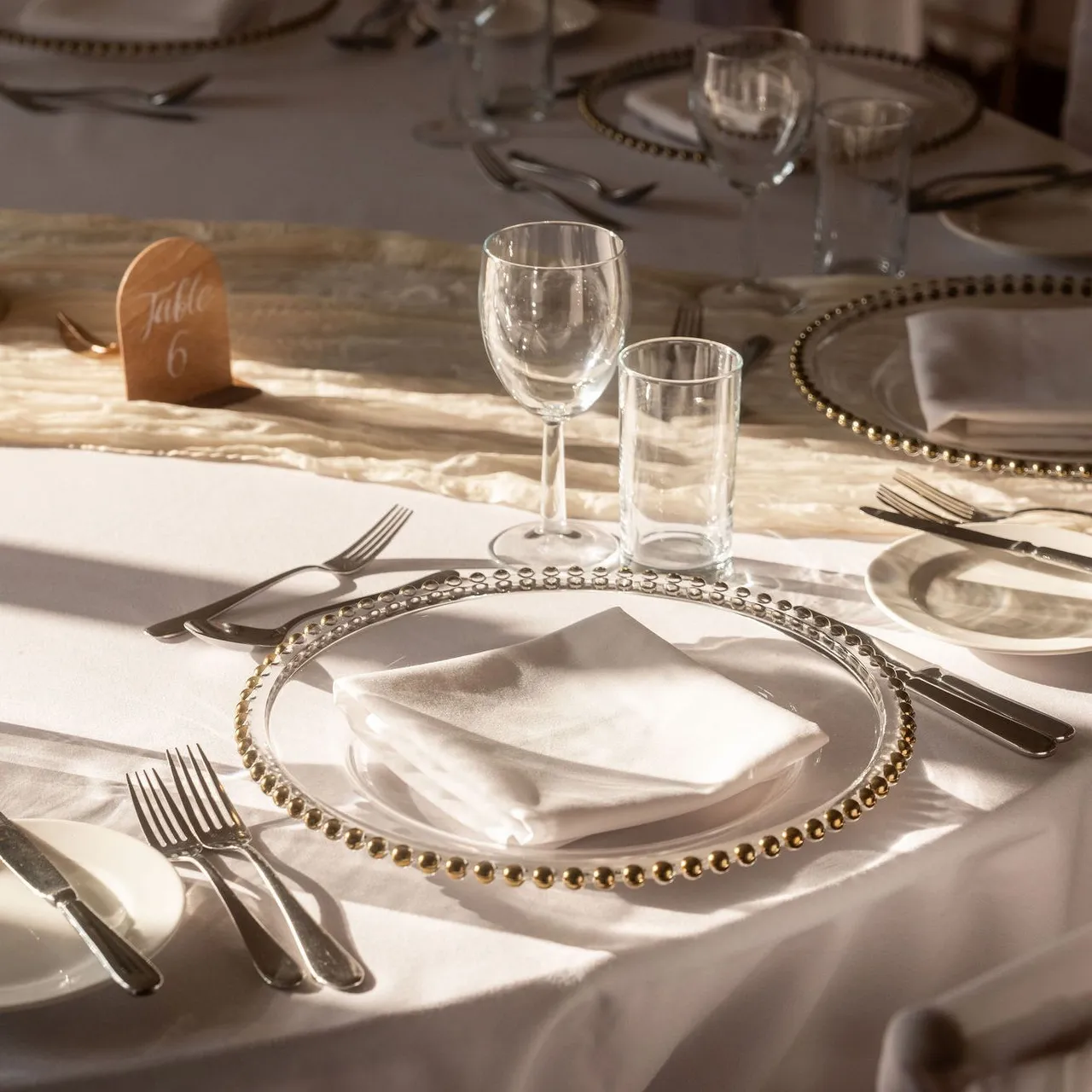 Elegant table setting with white linens, beaded charger plates, cutlery, glassware, and a table number sign.