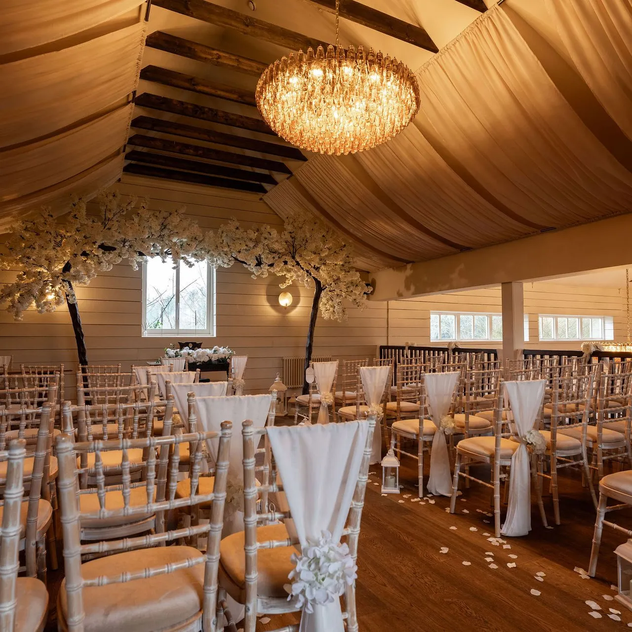 Elegant indoor wedding ceremony setup with rows of chairs, white drapes, flower arrangements, and chandeliers.
