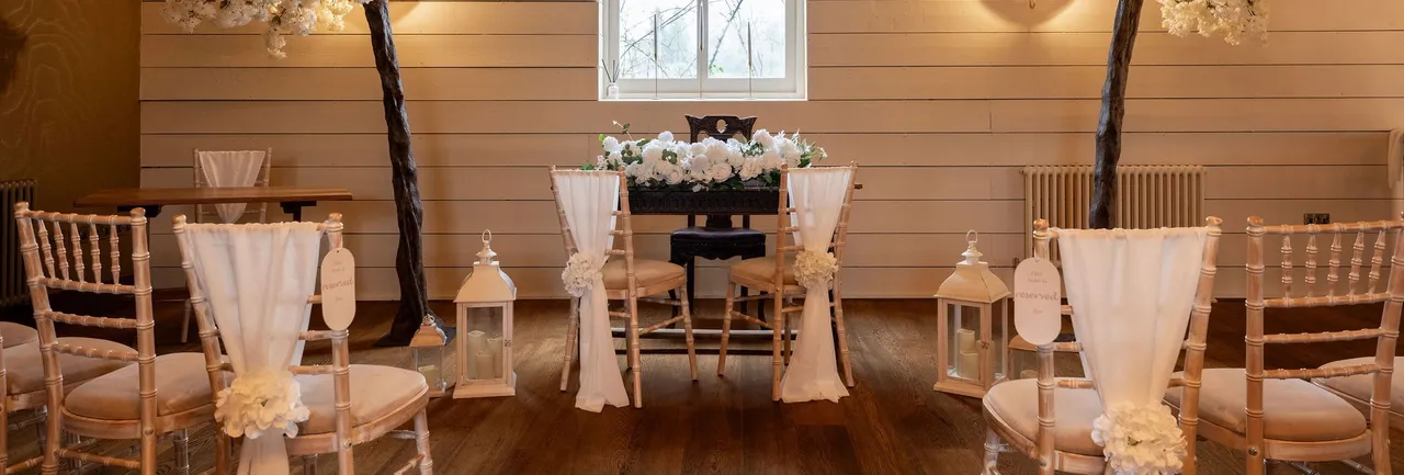 Wedding ceremony setup with white floral decorations, draped chairs, and lanterns in a rustic indoor venue.