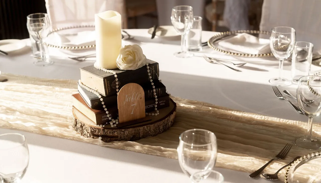 Elegant table centerpiece with stacked books, a candle, a white rose, pearl beads, and a wooden table number surrounded by tableware.